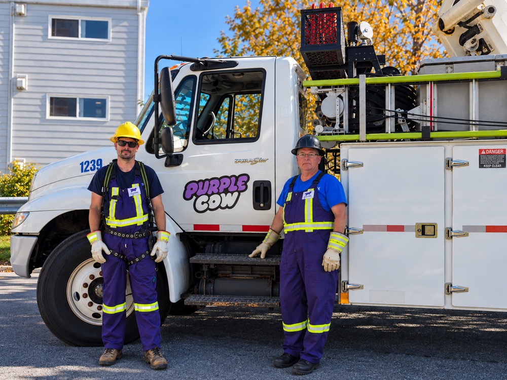 Two Purple Cow crew members standing next to a Purple Cow branded bucket truck. Both wear navy coveralls with high-visibility yellow safety stripes, hard hats, and work gloves.