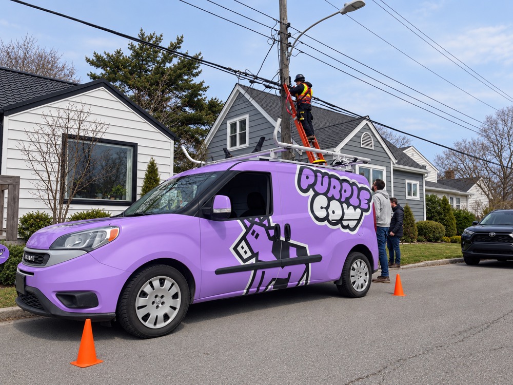 A Purple Cow branded van parked on a residential street in Fairview, with a Purple Cow crew member climbing a ladder to attach fibre to a utility pole. Two additional team members stand at the base of the pole.